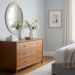 A beautifully styled dresser in a modern, light-filled bedroom, positioned against a wall opposite the bed. The dresser is a focal point, adorned with a decorative mirror, a ceramic vase with fresh flowers, and a stack of books, creating a peaceful and organized atmosphere.