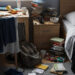 A realistic photograph of a very messy bedroom, featuring scattered clothes, books, and personal items on the floor and furniture, conveying the need for a thorough cleaning.