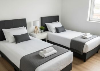 A high-quality, realistic photo of a modern twin bedroom featuring two parallel twin beds with dark grey headboards and minimalist bedding, separated by a nightstand. The room has white walls and a light wood floor, reflecting a clean and organized aesthetic for shared living spaces.