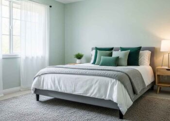 A serene and stylish bedroom with light sage green walls, a gray quilted comforter, and accent pillows in various shades of green. The room features a light wood nightstand with a lamp and a large window with sheer white curtains.