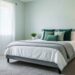A serene and stylish bedroom with light sage green walls, a gray quilted comforter, and accent pillows in various shades of green. The room features a light wood nightstand with a lamp and a large window with sheer white curtains.